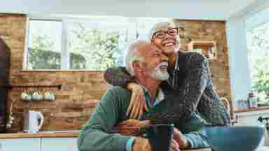 A happy elderly couple sitting at a table in the kitchen and hugging.