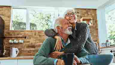 A happy elderly couple sitting at a table in the kitchen and hugging.