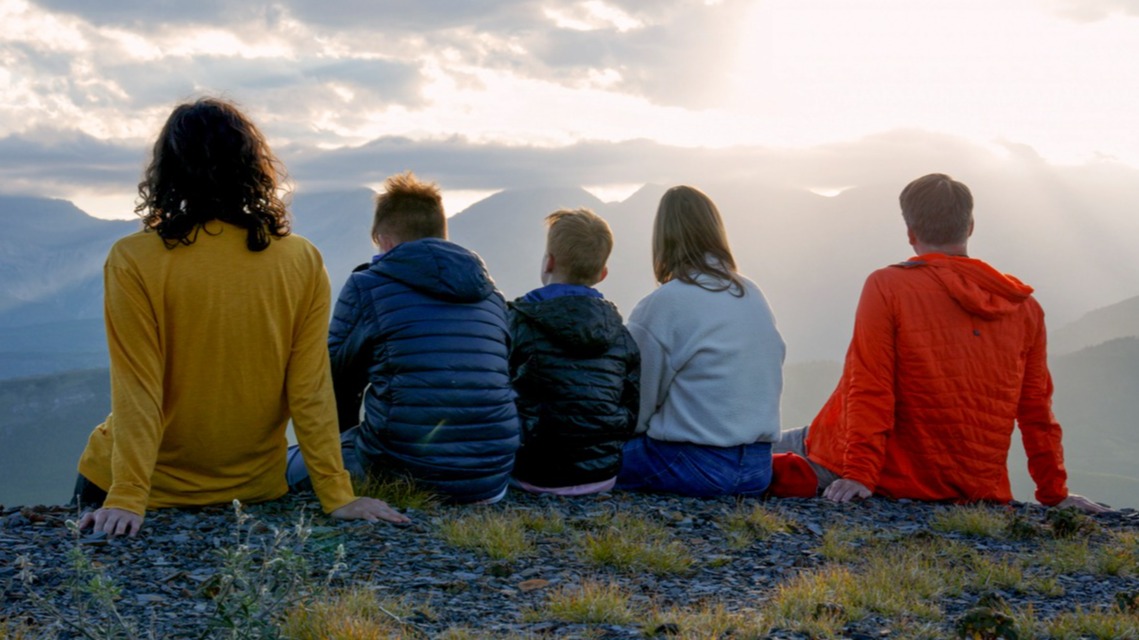 Five people sit side by side on a rocky mountain summit, backs to camera, gazing at a glowing sunset over misty mountain ranges.