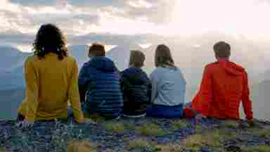 Five people sit side by side on a rocky mountain summit, backs to camera, gazing at a glowing sunset over misty mountain ranges.