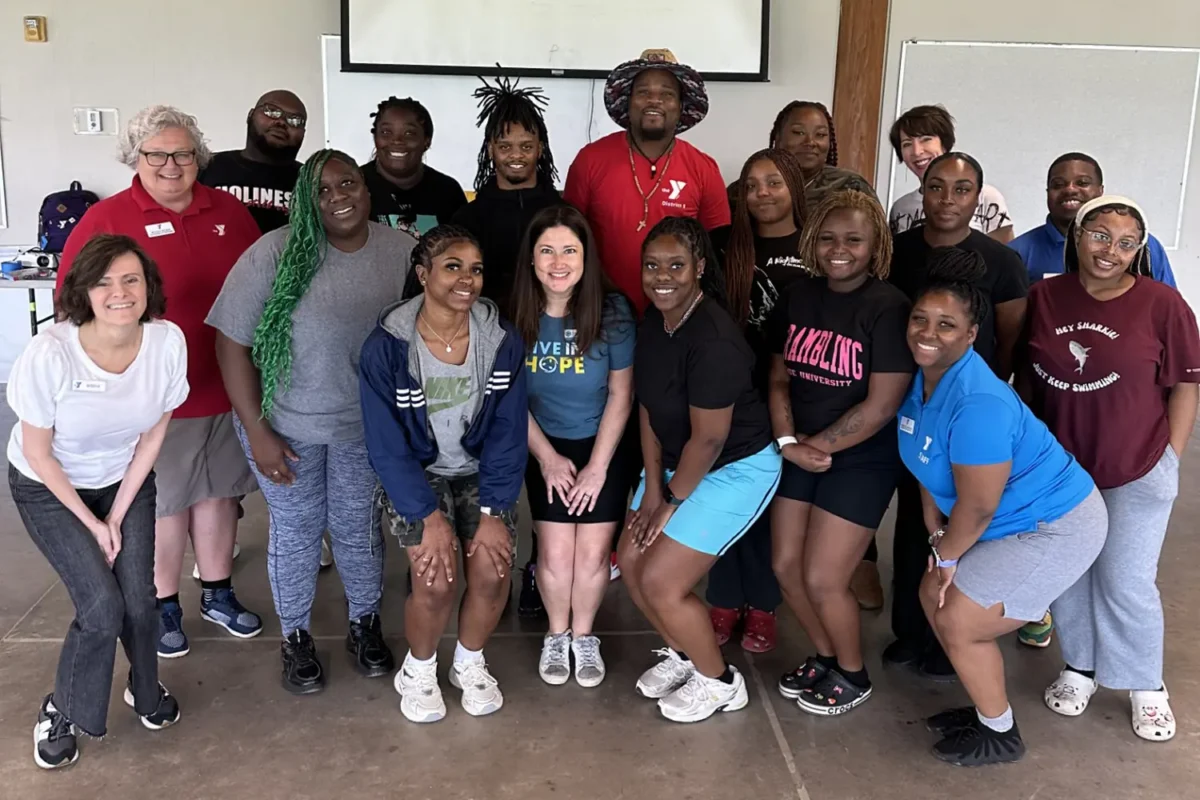 A diverse group of smiling adults poses together indoors, some wearing YMCA shirts, in a casual group photo.