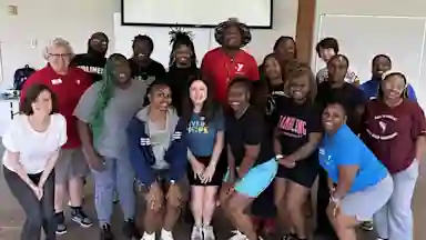 A diverse group of smiling adults poses together indoors, some wearing YMCA shirts, in a casual group photo.