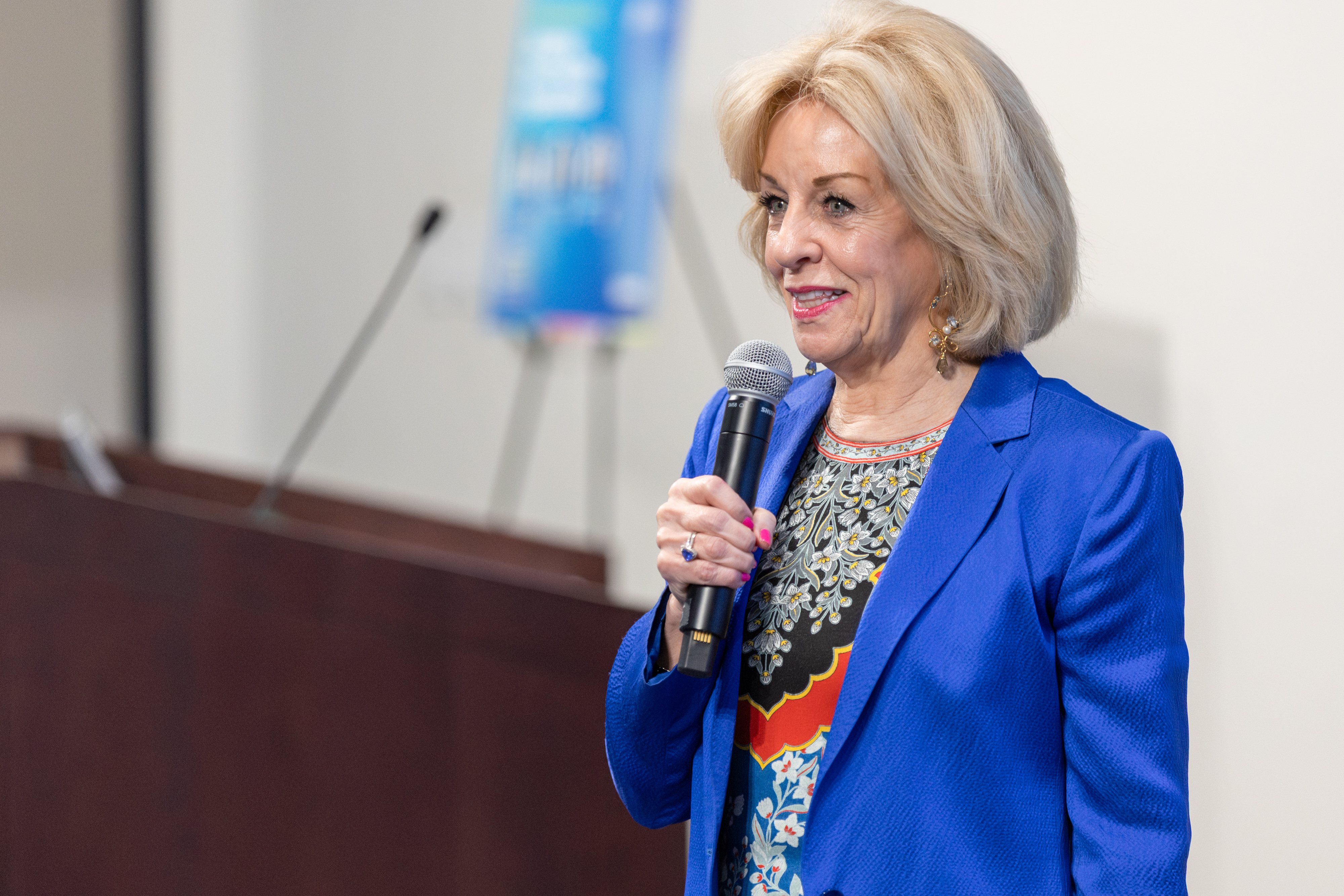 Dr. Sandi Chapman in a blue blazer and floral top holds a microphone while speaking at an event near a wooden podium.