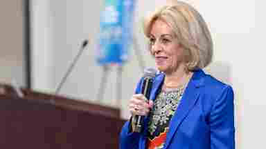Dr. Sandi Chapman in a blue blazer and floral top holds a microphone while speaking at an event near a wooden podium.