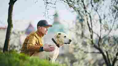 A relaxed young man is with a dog on the grass in a public park located in Prague, Czech Republic. He is happy and content while being in nature.