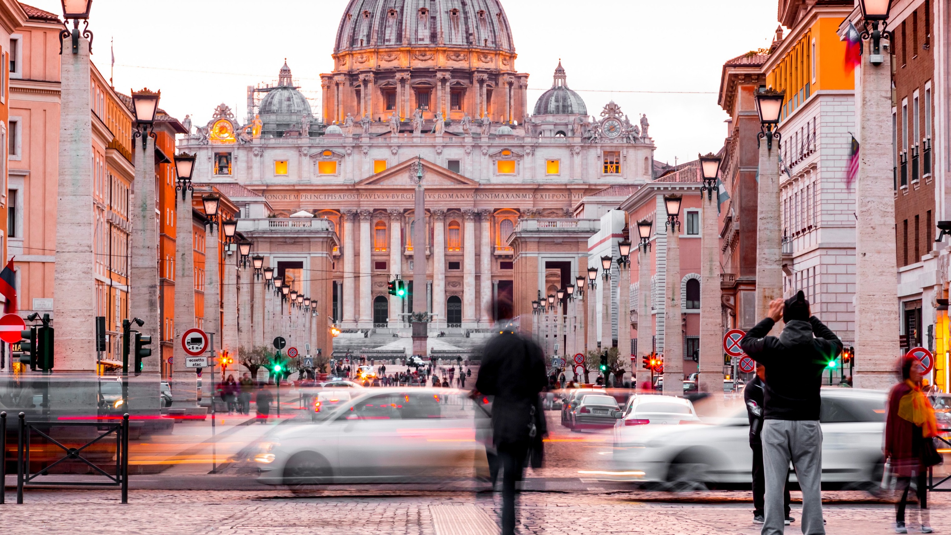 St. Peter's Basilica at dusk, viewed from a busy Rome street with blurred pedestrians and moving cars in the foreground.