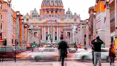 St. Peter's Basilica at dusk, viewed from a busy Rome street with blurred pedestrians and moving cars in the foreground.