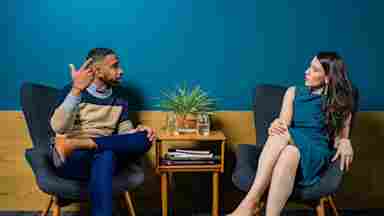 Women wearing a blue dress, sitting on a dress talking to a man of color in a colorful sweater and jeans.