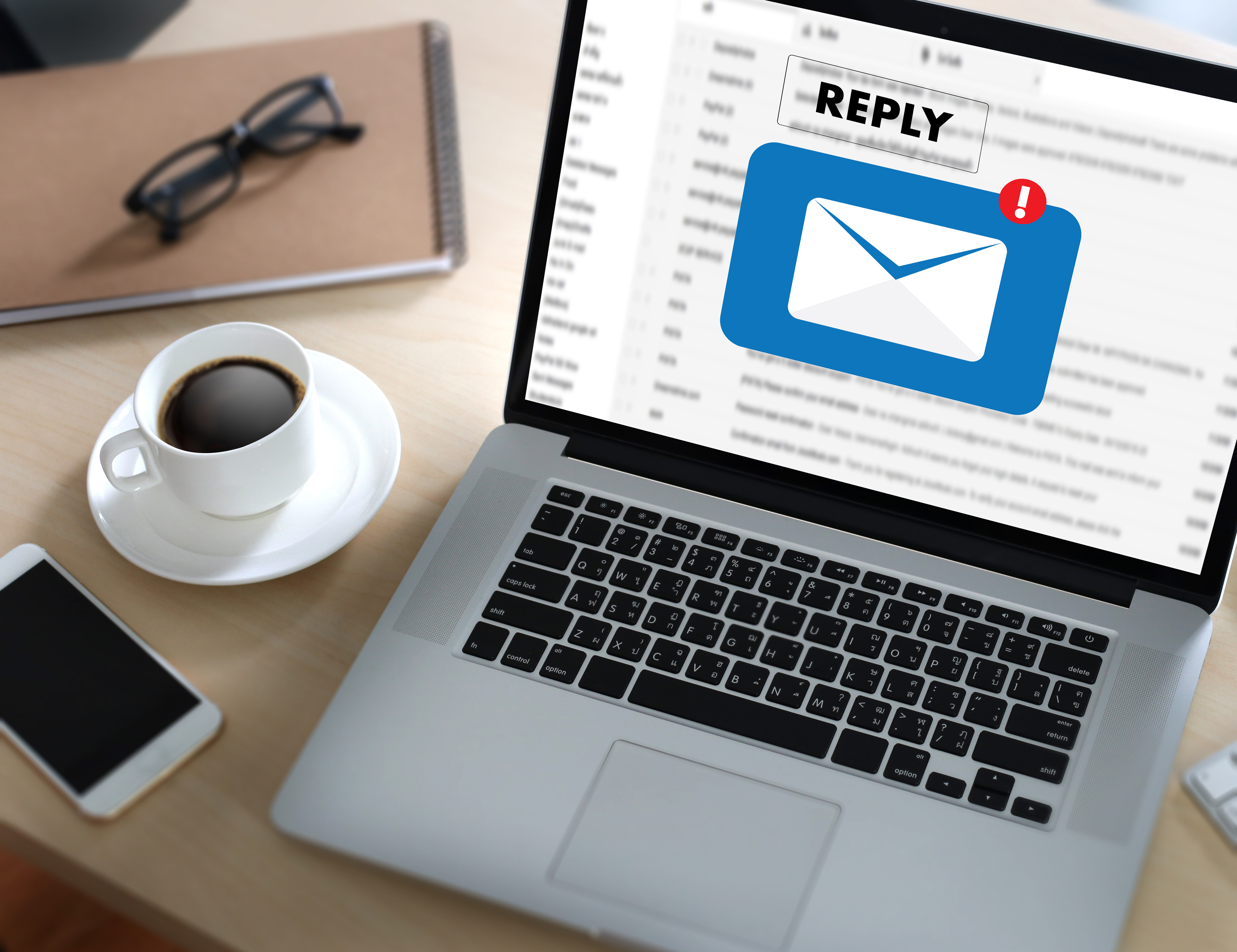 Laptop on a desk displaying an email reply notification icon, with a coffee cup, glasses, notebook, and smartphone nearby.