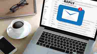Laptop on a desk displaying an email reply notification icon, with a coffee cup, glasses, notebook, and smartphone nearby.