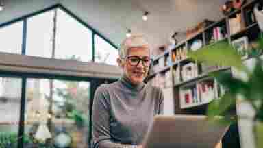 An elderly woman is typing on a laptop surrounded by books and plants.