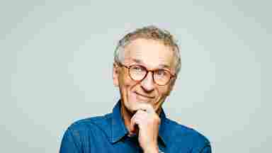 A middle-aged man with glasses and a blue shirt against a light gray background; his hand is on his chin and he is smiling thoughtfully.