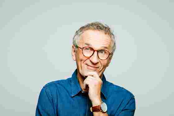 A middle-aged man with glasses and a blue shirt against a light gray background; his hand is on his chin and he is smiling thoughtfully.