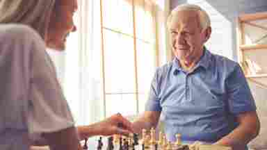 A smiling woman is playing chess with a smiling elderly male in a house setting. IStock# 619393614.