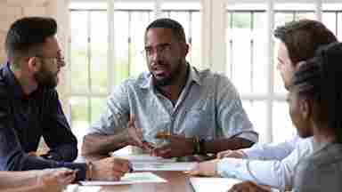 A man is talking to his colleagues at work, as they gather around a table. Black male.
