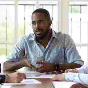 A man is talking to his colleagues at work, as they gather around a table. Black male.
