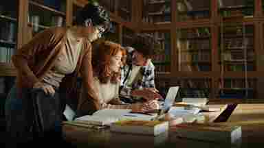 A group of high school students work together on a laptop in the library.