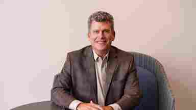 Stephen White in a brown sports coat is sitting down with a plain background, landscape. Chief Operating Officer