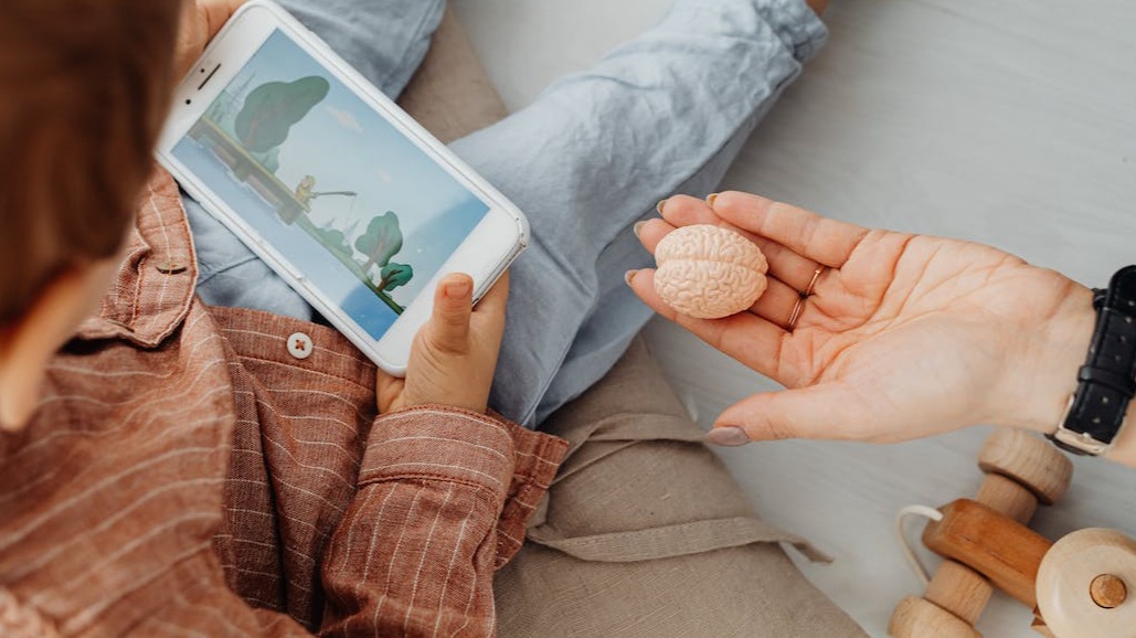 Child playing with their phone while a hand presents a brain-shaped eraser to them