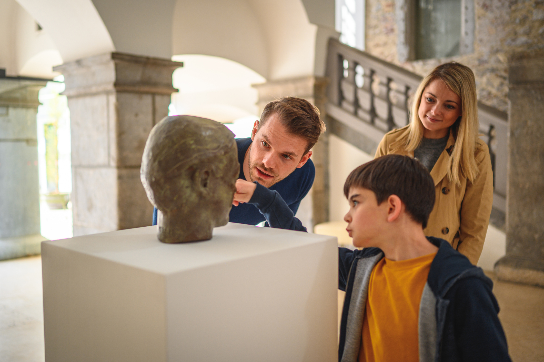 A curious child takes a closeup look at an antique bronze head in a museum, as two adults share the child's interest.