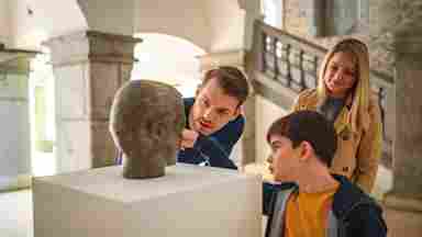 A curious child takes a closeup look at an antique bronze head in a museum, as two adults share the child's interest.