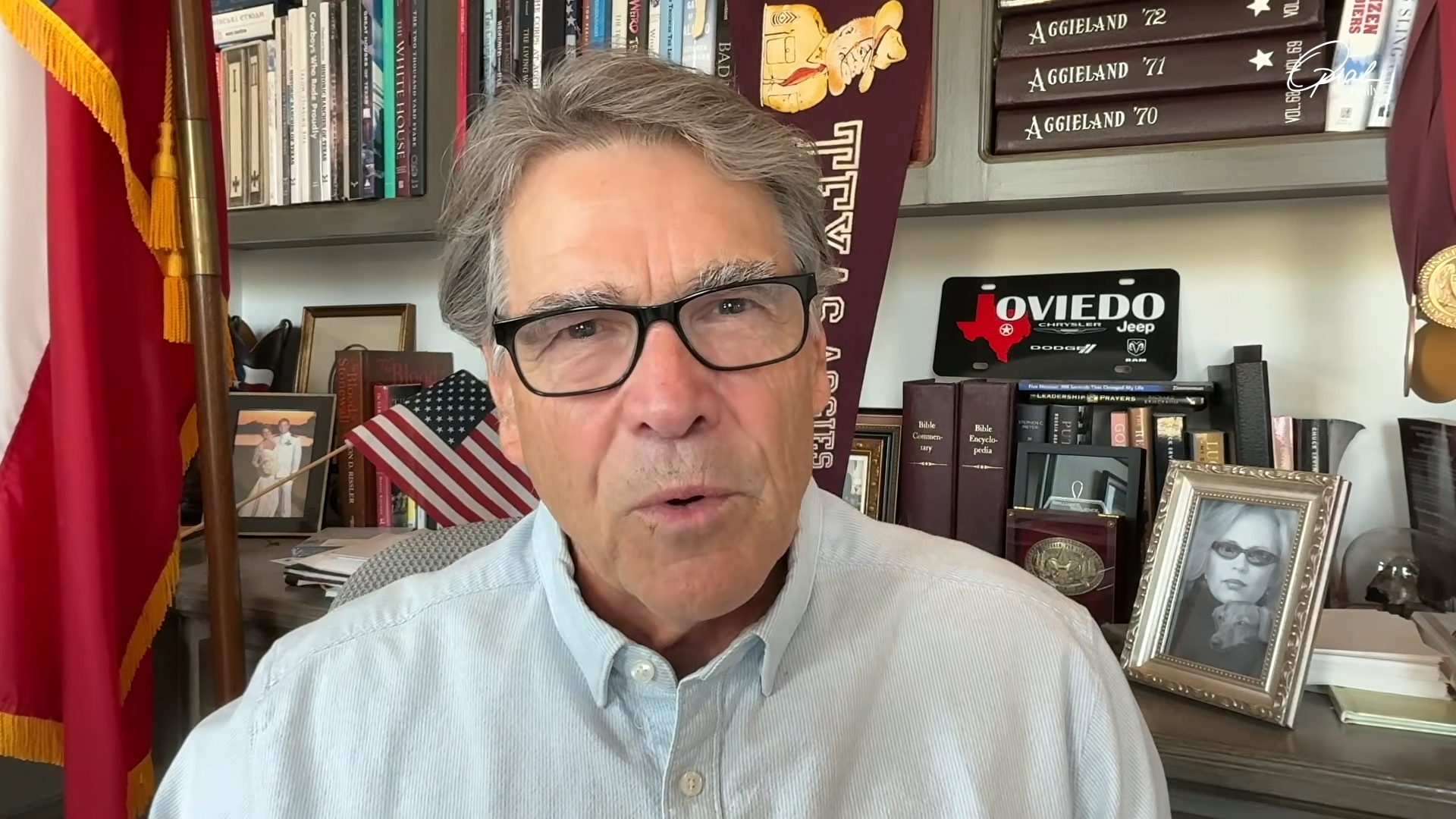 A man with gray hair and black glasses speaks in an office with books, flags, and Aggieland memorabilia in the background.