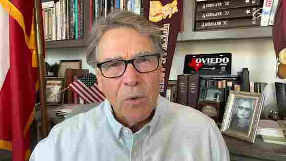 A man with gray hair and black glasses speaks in an office with books, flags, and Aggieland memorabilia in the background.