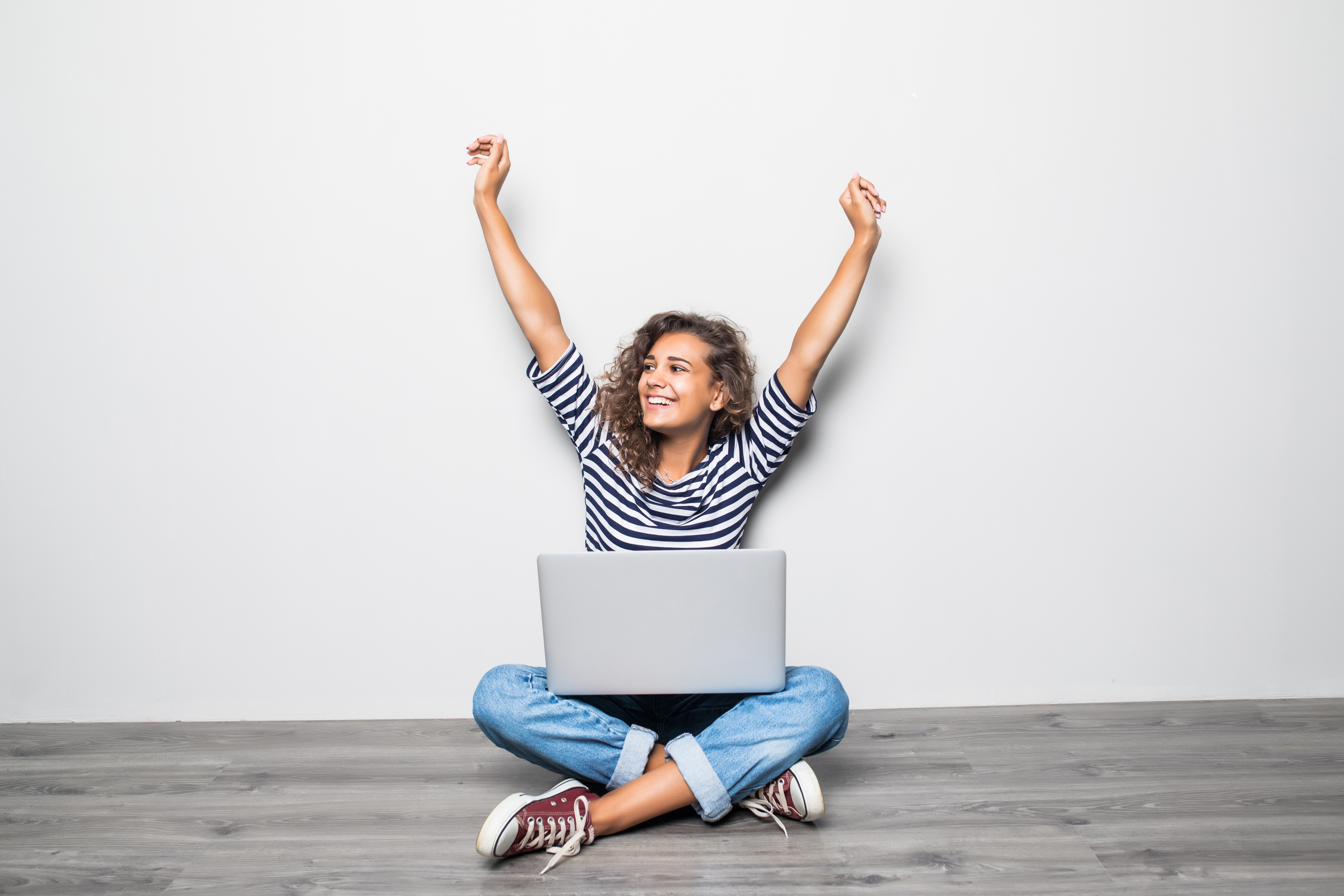 Young woman sitting cross-legged on floor with laptop, arms raised in celebration, wearing striped shirt and jeans, smiling.