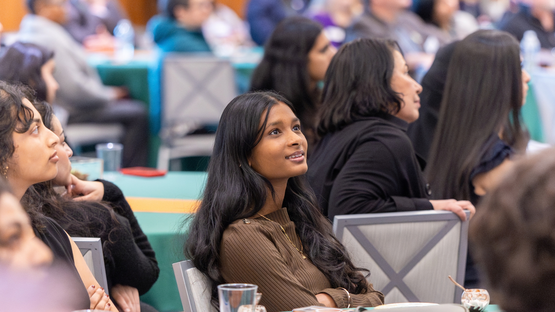 A young UT Dallas student with long dark hair and a brown top looks up attentively at NextGen Day during BrainHealth Week, seated at a teal-covered table.