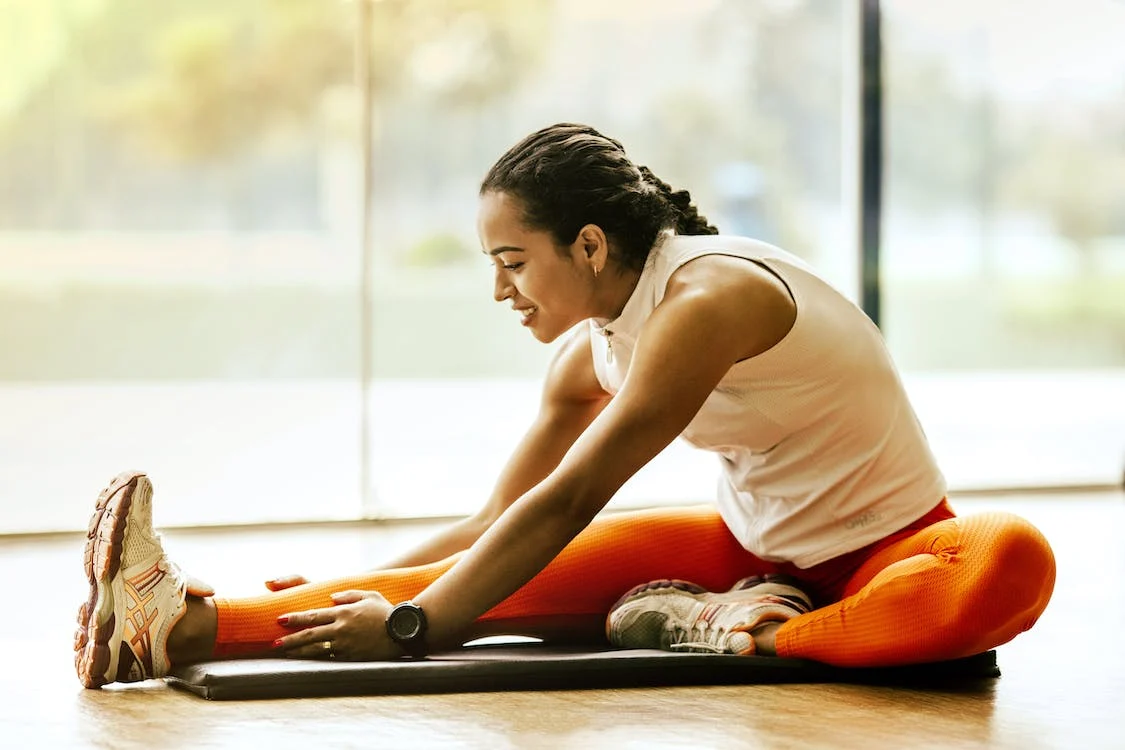 A woman of color stretching by a sunlit window before yoga class.