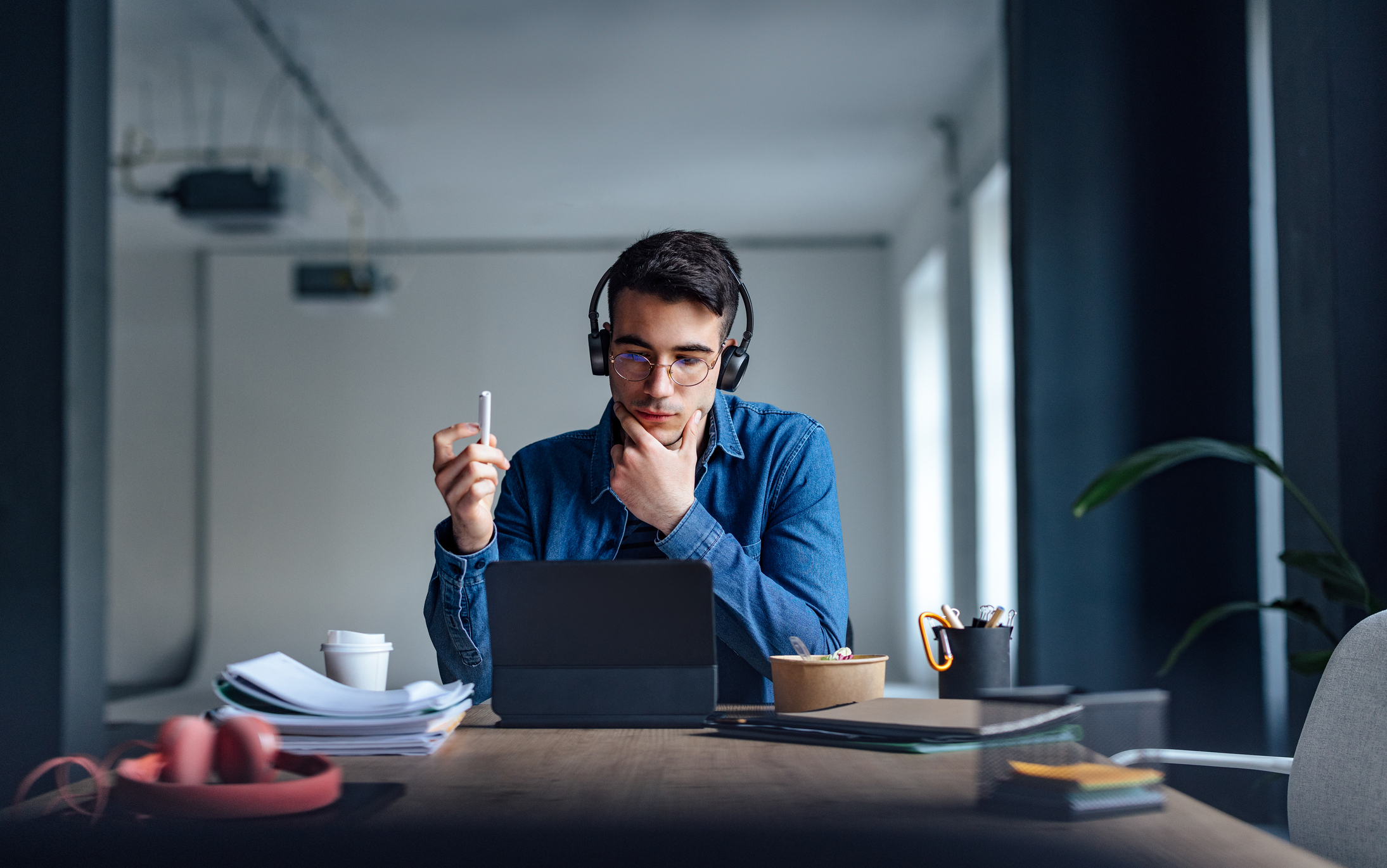 Young man wearing headphones and glasses sits at a cluttered desk, thoughtfully looking at a tablet while holding a pen.