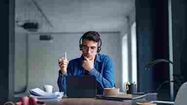 Young man wearing headphones and glasses sits at a cluttered desk, thoughtfully looking at a tablet while holding a pen.