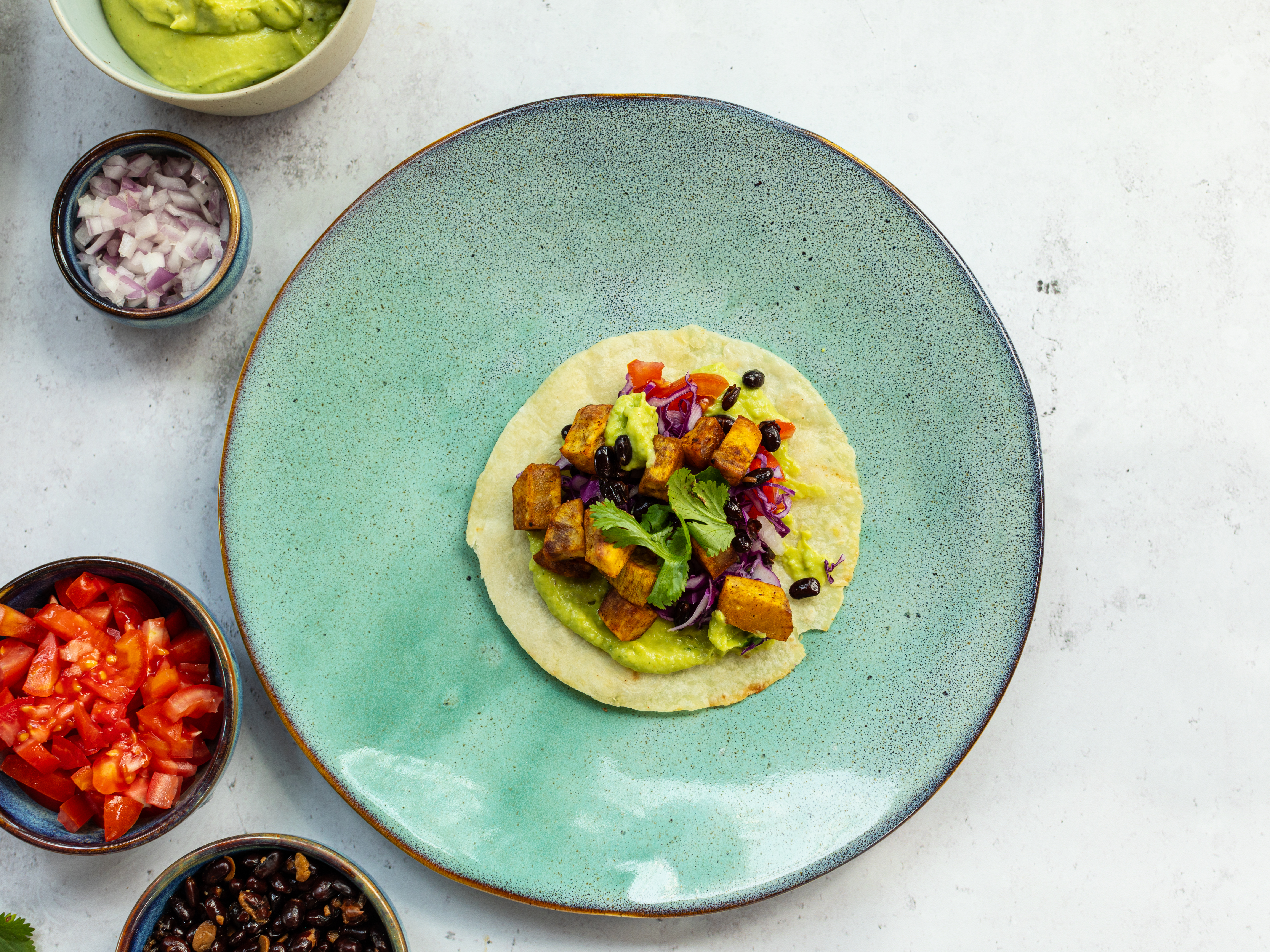 Schritt 4: Die Tortillas mit der pürierten Avocado-Creme bestreichen und mit den gebackenen Süsskartoffeln, gewürfelter Zwiebel, dünn geschnittenem Rotkohl, Bohnen, Tomaten und frischem Koriander füllen
