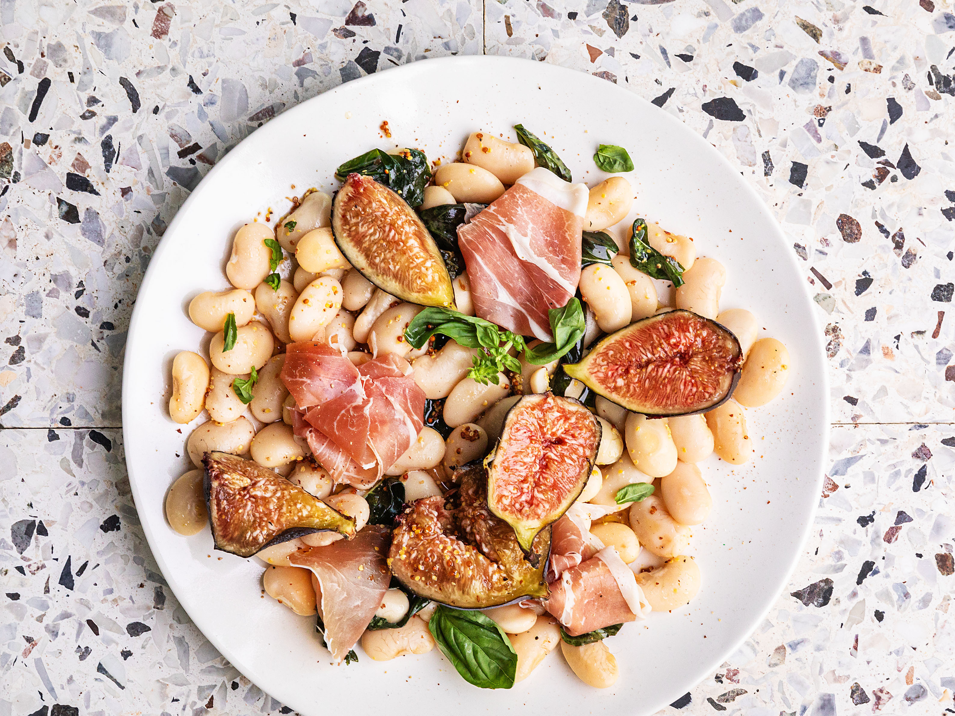 Schritt 4: Die fertigen Bohnen auf einer Servierplatte anrichten, den Schinken und die Feigen dazugeben und mit etwas Olivenöl beträufeln
