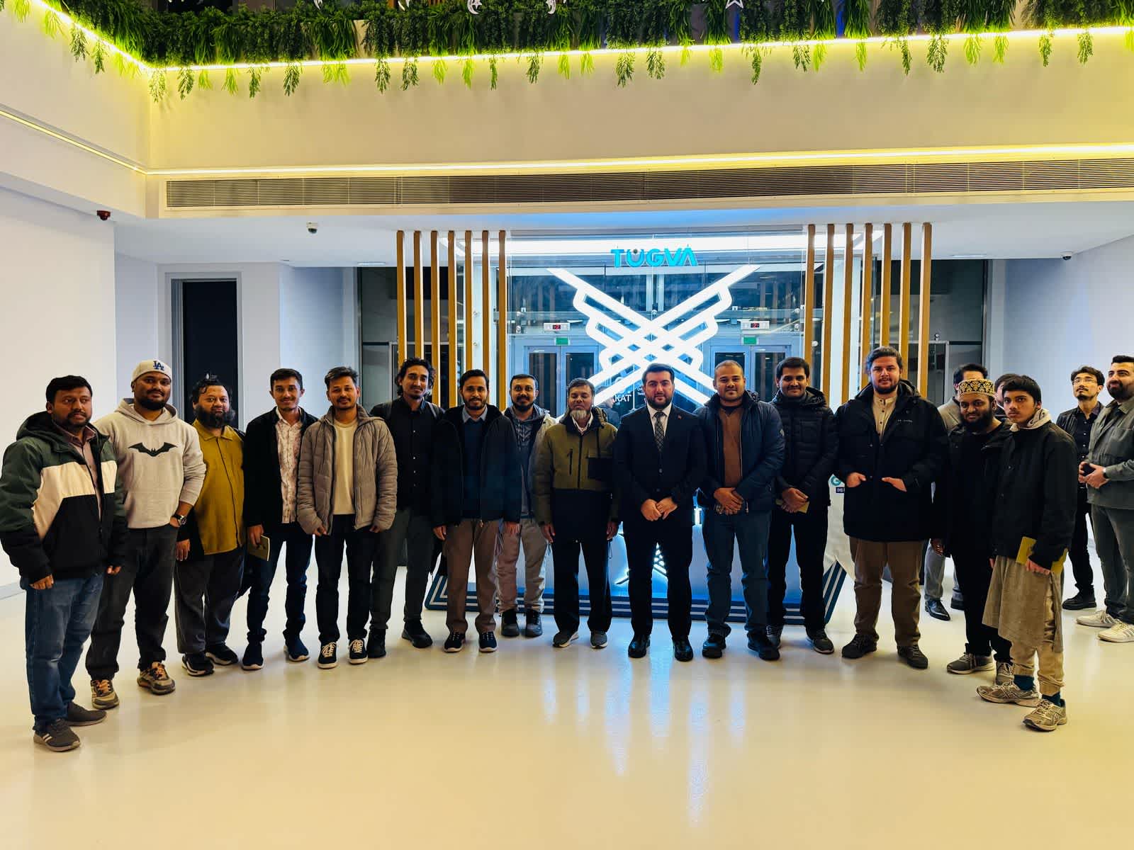 A group photo of the Runner Bangladesh Development Society delegation in front of the TÜGVA logo, showcasing the official visit and diplomatic engagement between the two organizations.