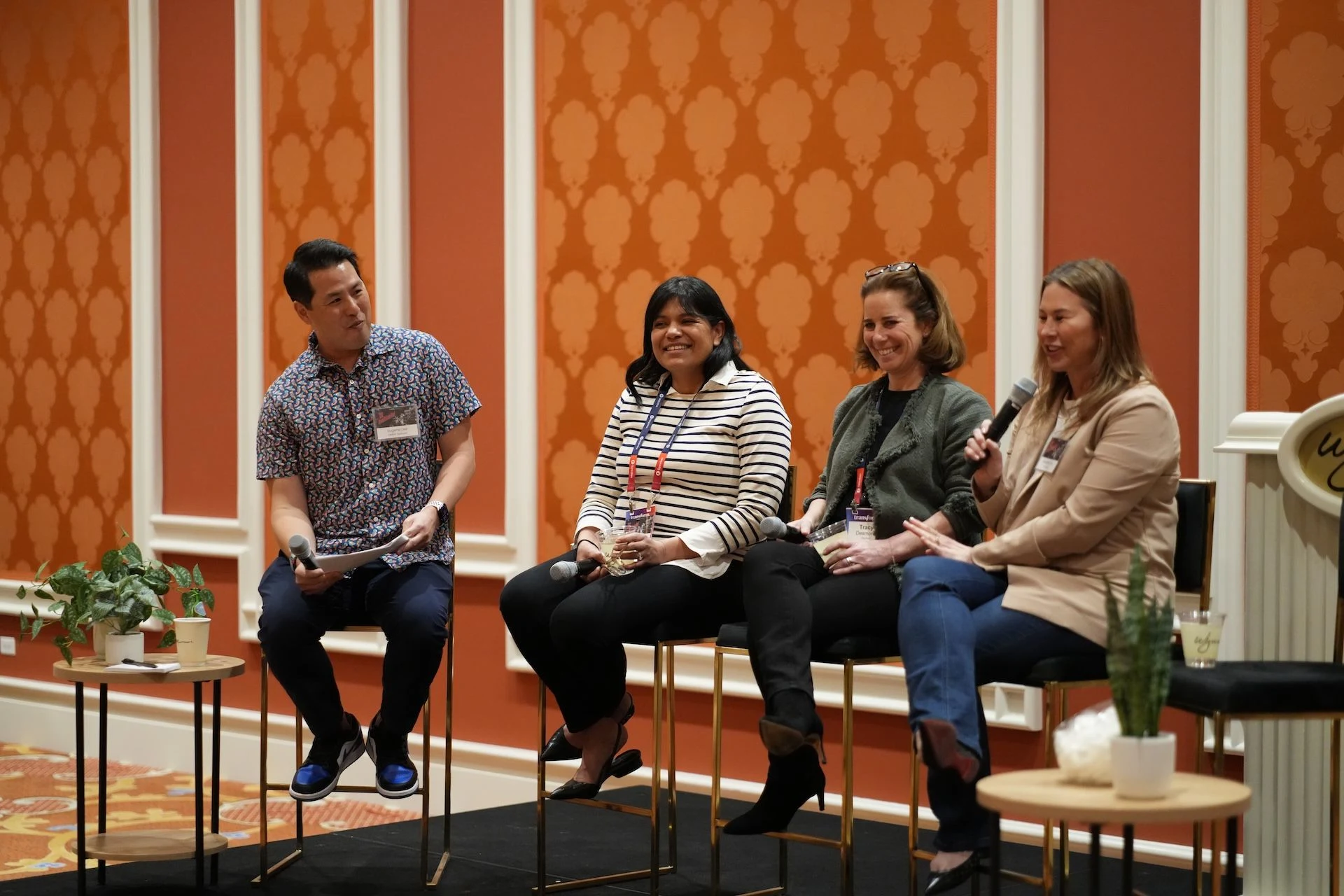 A picture of four people speaking on a panel at a conference center, with one moderator and three panelists. They're seated on stool-height chairs.