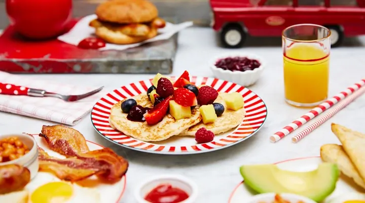 Plate of pancakes topped with fruit next to a glass of orange juice and paper straws