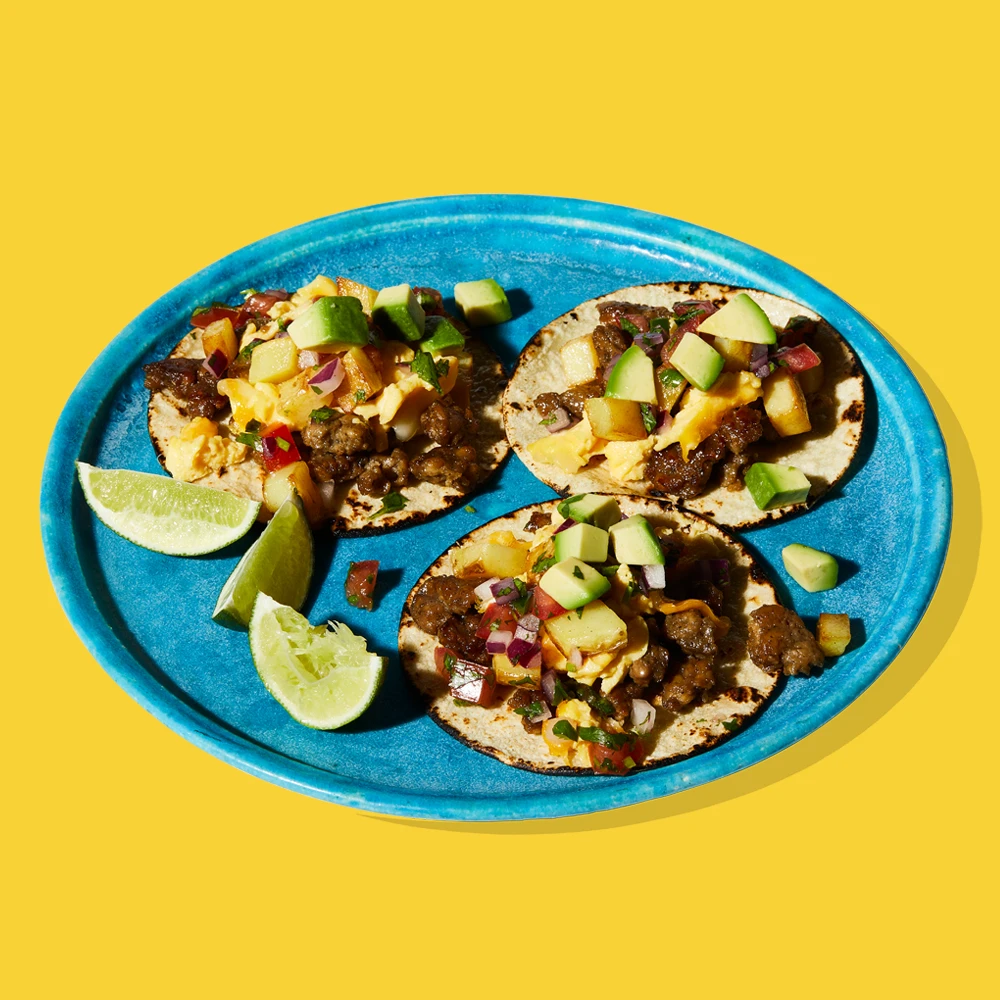 Three Impossible Sausage breakfast tacos on a blue plate, each piled high with plant-based meat, potatoes, scrambled eggs, chopped avocado, and salsa, served with wedges of fresh lime. 