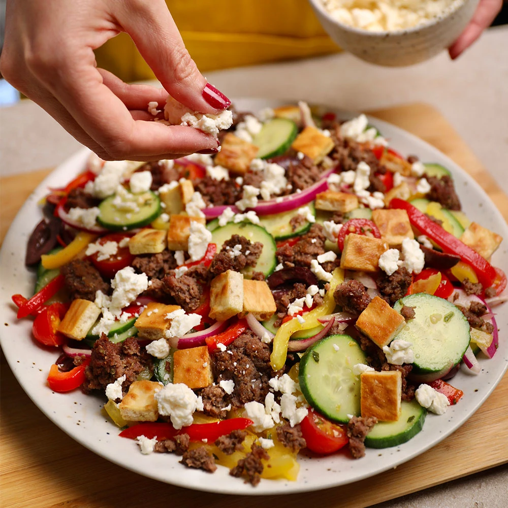An Impossible Greek salad featuring seasoned Impossible Beef Lite Made From Plants, cucumber, cherry tomatoes, kalamta olives, feta cheese, and pita croutons. 
