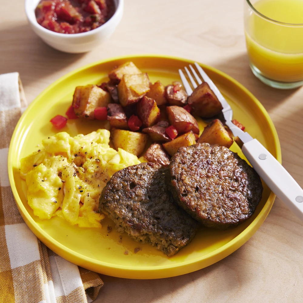 A complete breakfast plate with two Impossible™ Sausage Patties, scrambled eggs and roasted potatoes with red peppers, alongside a small dish of salsa and a glass of orang juice. 