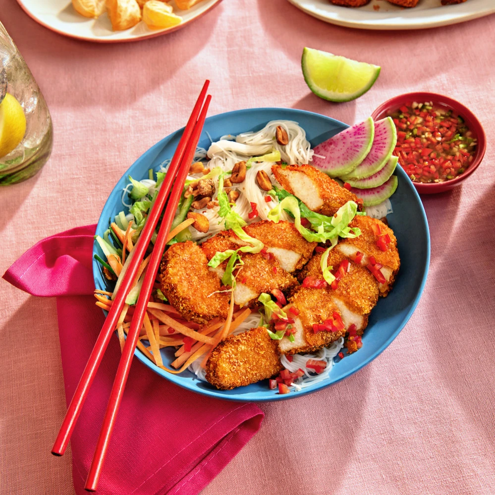 A noodle bowl featuring Impossible Chicken Tenders Made From Plants, vermicelli noodles, shaved watermelon radish, thinly sliced carrot and romaine lettuce, served with a spicy sweet dressing, and fresh lime wedges. 