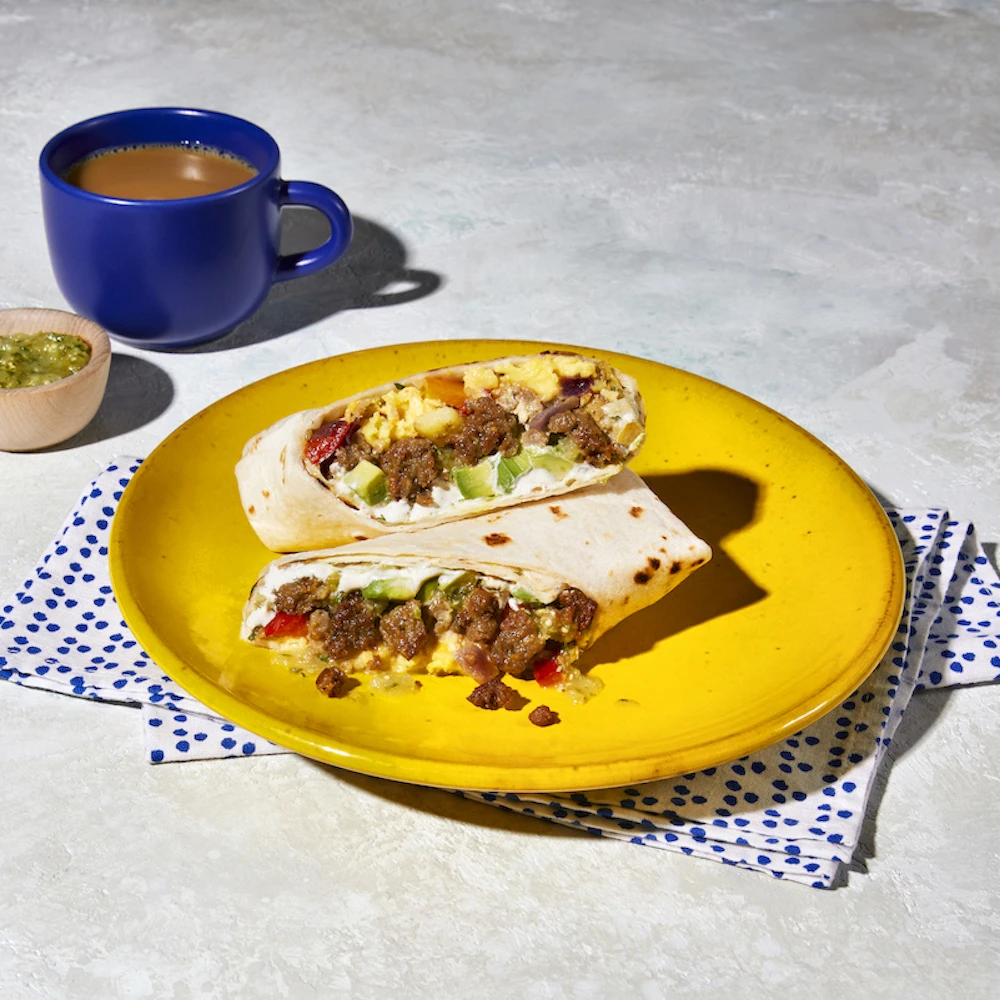 A cut open Impossible™ Sausage Breakfast Burrito with scrambled egg, Impossible Sausage, avocado, salsa, and cilantro lime crema, on a yellow plate with a napkin and a cup of coffee and side of tomatillo salsa. 