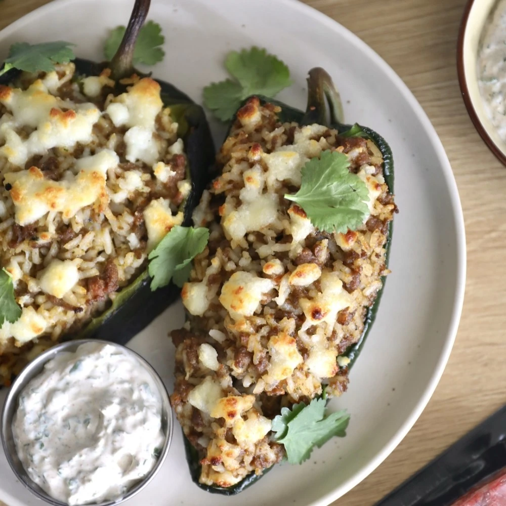 Two Impossible Beef Sofrito stuffed poblano peppers on a plate with browned, melted cheese, cilantro leaves and chipotle lime crema. 