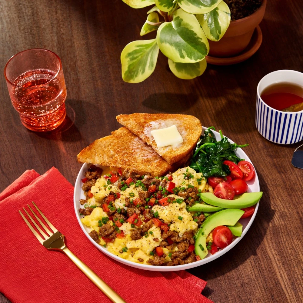 A delicious breakfast plate featuring a scramble of Impossible™ Savory Ground Sausage, buttered toast, sauteed spinach, and fresh sliced cherry tomatoes and avocado. 