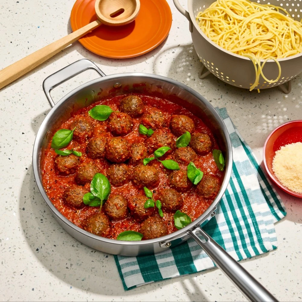 A large pot of Impossible Italian Meatballs garnished with cracked pepper and fresh basil, a colander of spaghetti waiting to be mixed in and finished with parmesan cheese. 