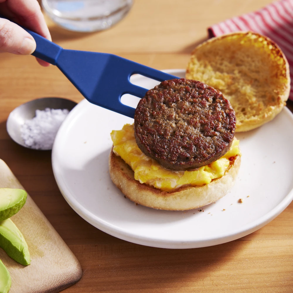 A perfectly golden Impossible™ Sausage Patty being placed on fluffy scrambled eggs on a buttered english muffin, with flakey salt and sliced avocado to complete the sandwich. 