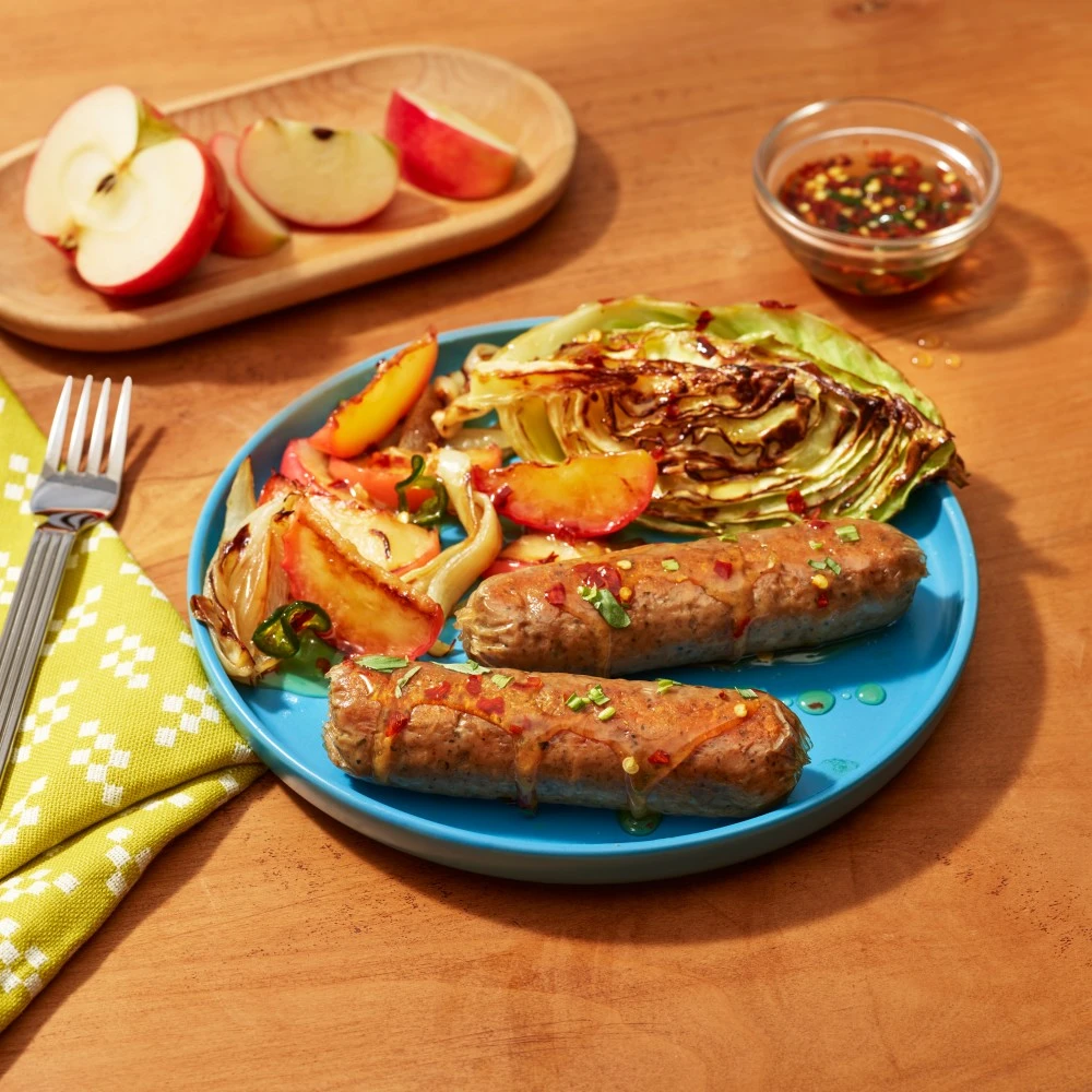 Two Impossible Bratwurst Sausage Links dripping with honey infused with jalapenos and chili flakes, with caramelized cabbage steak, and juicy, roasted apples and onions, on a blue plate. 