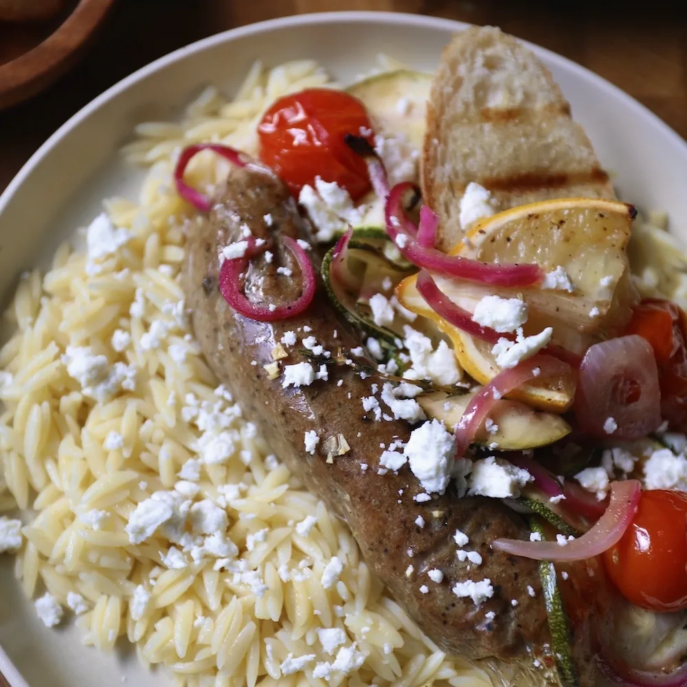 An Impossible Italian Sausage Link Made From Plants with roasted tomatoes, red onions, and yellow summer squash, sprinkled with feta cheese and served over orzo pasta. 