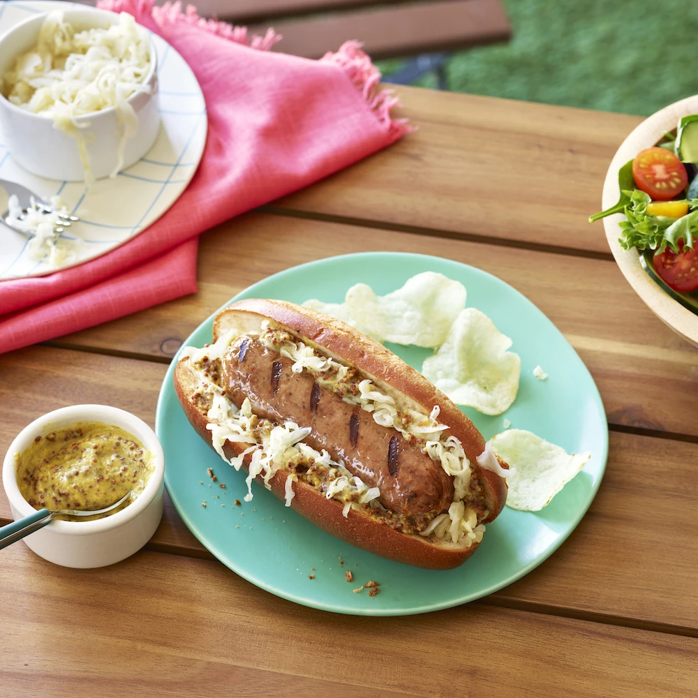 Impossible™ Grilled Bratwurst Sausage Link on a bun with whole grain mustard and sauerkraut with potato chips and a fresh side salad, all on a picnic table outside. 