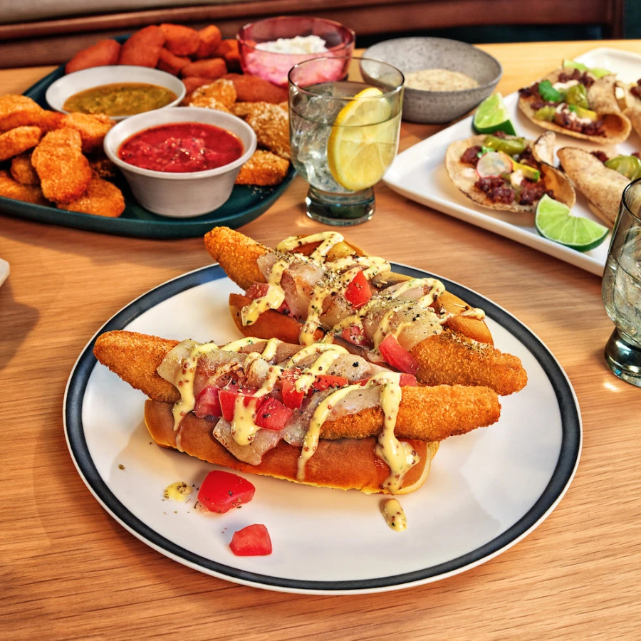 Impossible Bird Dog Sandwich featuring Impossible Chicken Tenders Made From Plants, served in a hot dog bun with melted cheese and diced tomatoes, drizzled with whole grain honey mustard. 